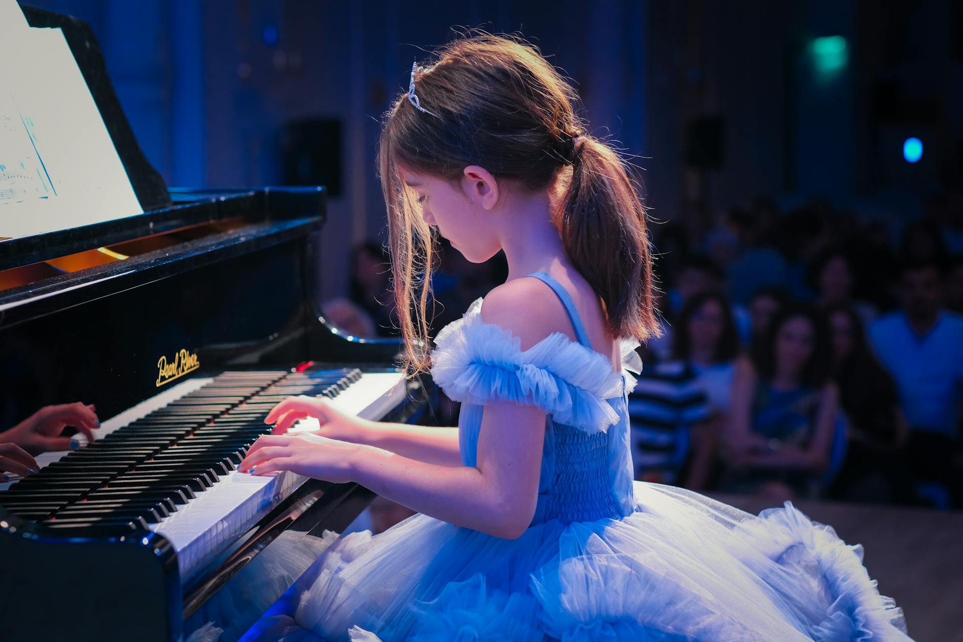 Dziewczynka w niebieskiej sukience gra na fortepianie przed publicznością